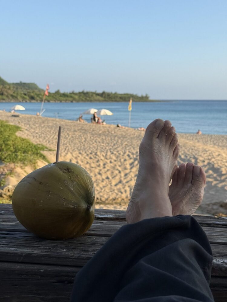 A fresh coconut beside sandy feet resting on a wooden platform overlooking the beach and sea in Kenting.
